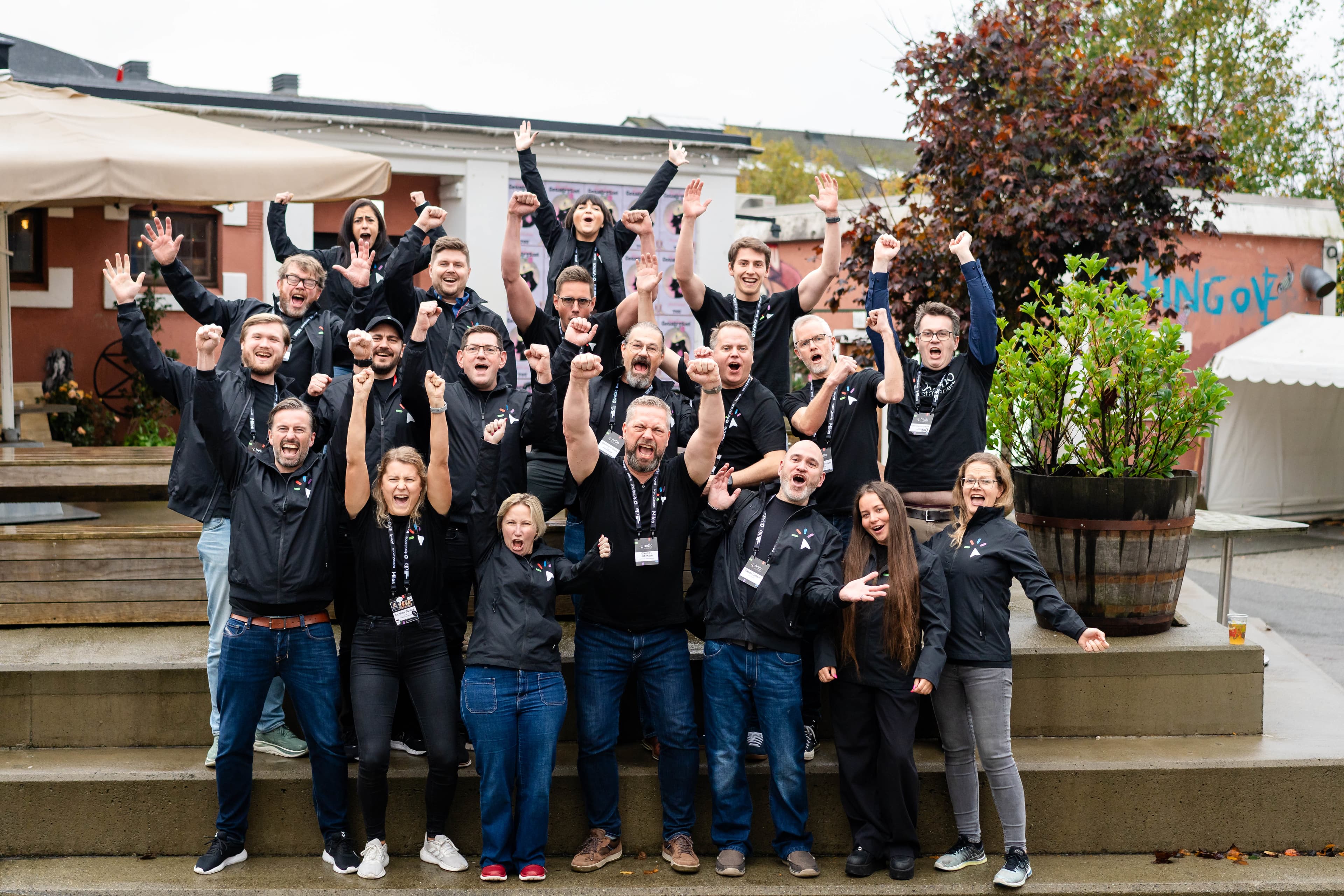 Hello Stavanger organizers — a diverse team cheering together on outdoor steps in matching black jackets.